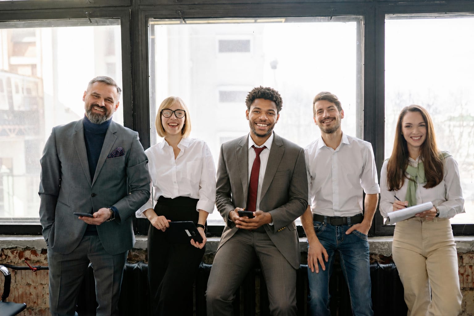 Five colleagues standing in a row in front of tall windows, smiling