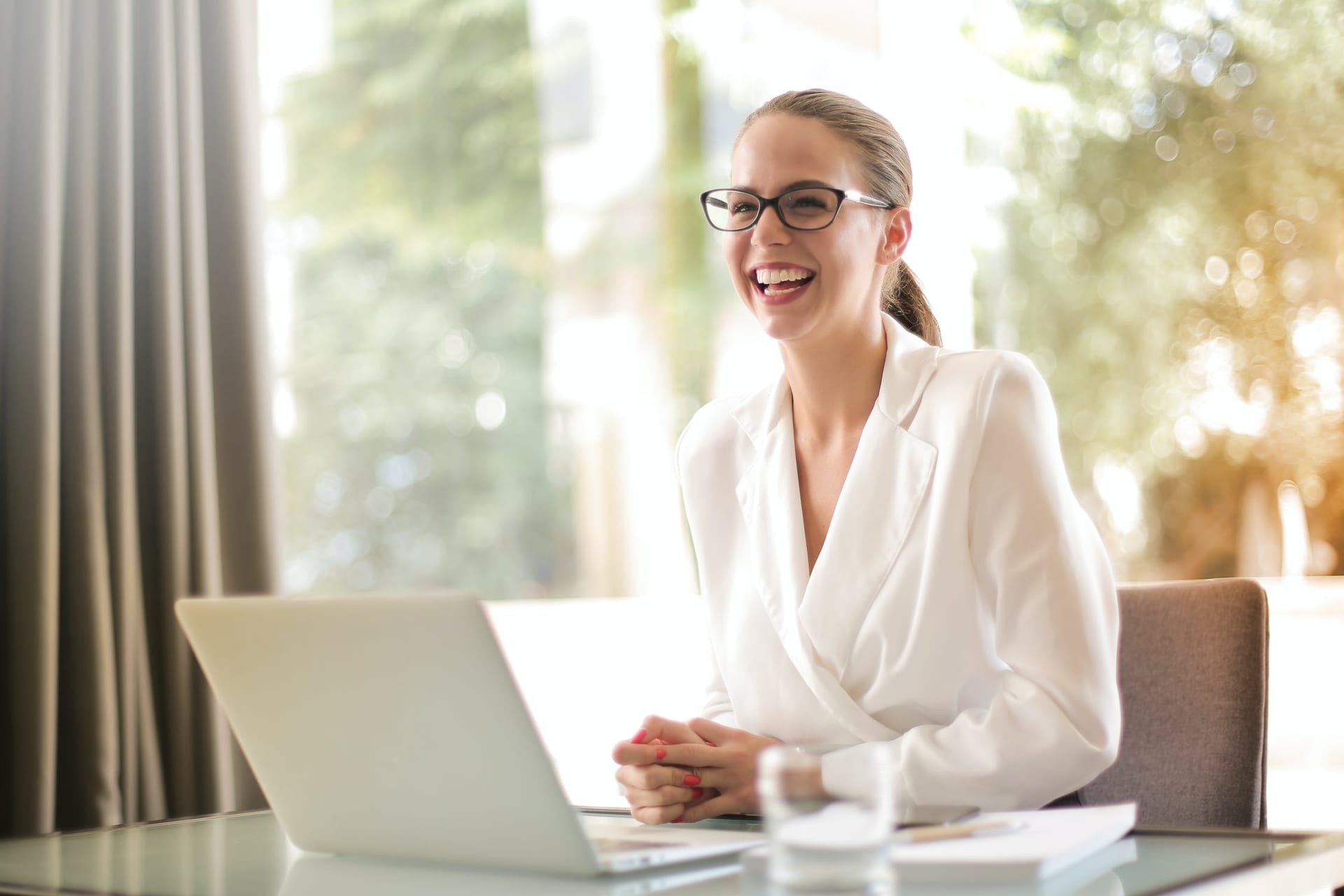 A woman in glasses laughing at a desk with a laptop open in front of her