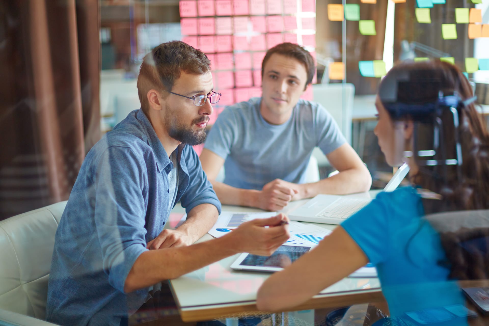 Three people in discussion at a table in front of a wall of sticky notes